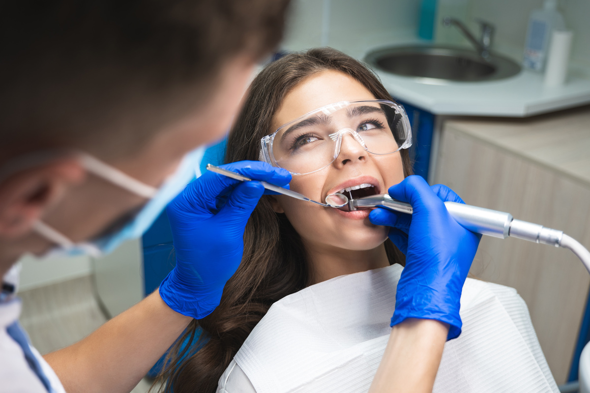 dentist in mask filling the patient's root canal while she is lying on dental chair in safety glasses under the medical lamp in clinic.