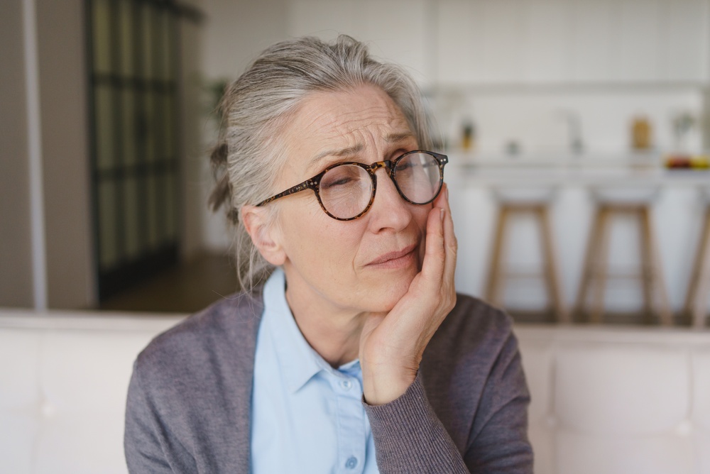 Older woman pressing a hand to her cheek from persistent tooth and gum pain at home.