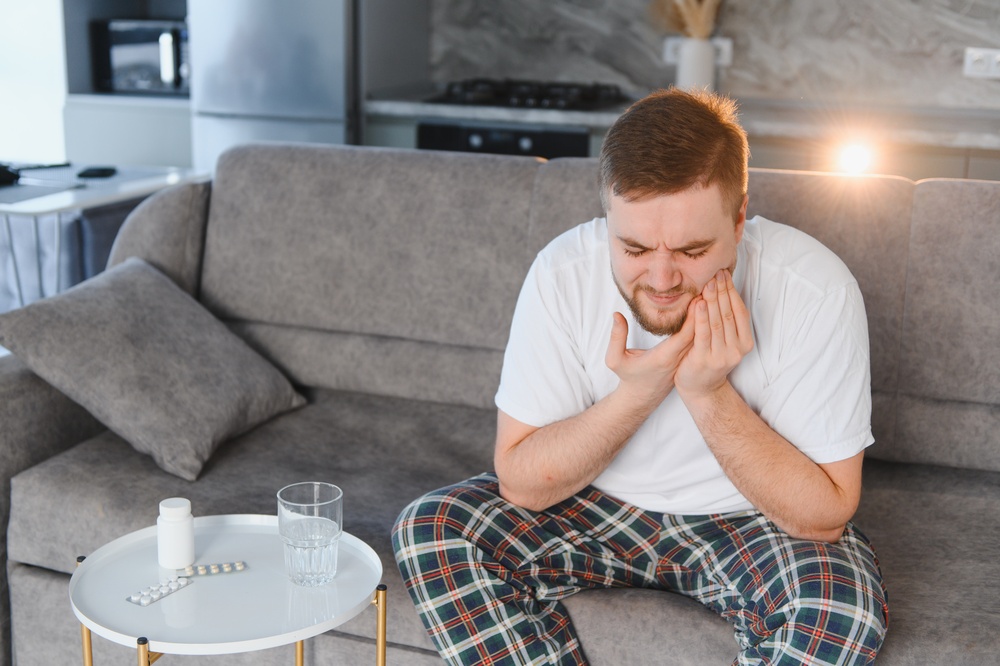 Man holding his jaw at home beside pain relievers and a glass of water while coping with a toothache