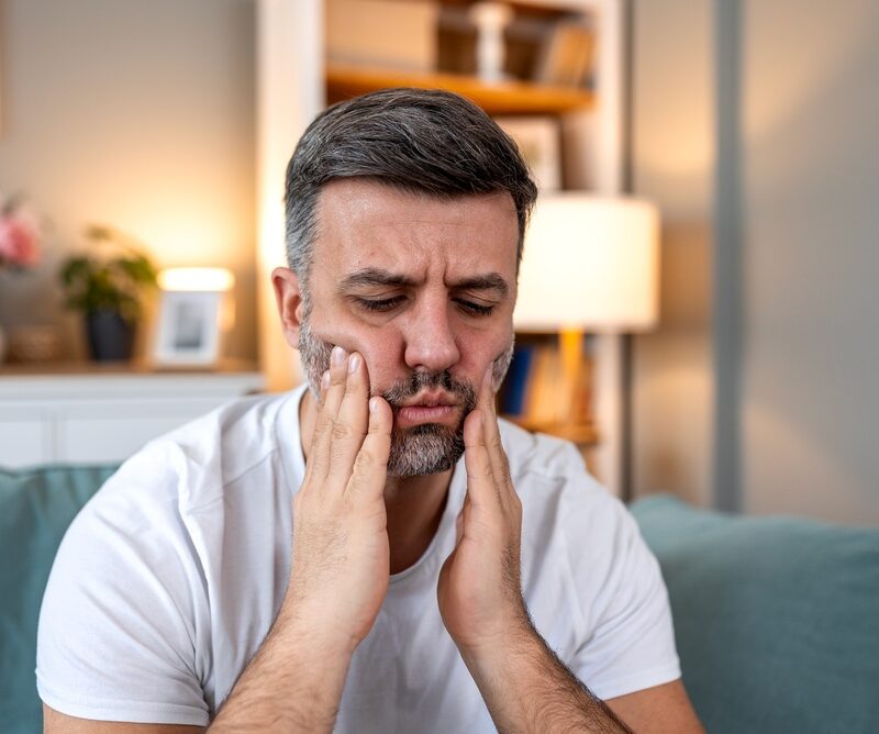 Man wincing in pain and holding both hands to his jaw, showing signs of a dental emergency that need immediate care