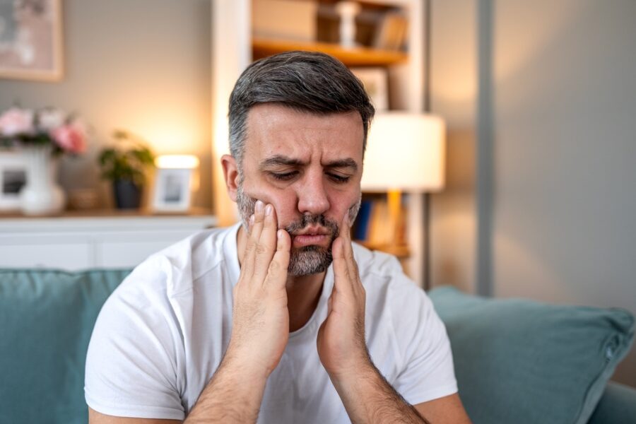 Man wincing in pain and holding both hands to his jaw, showing signs of a dental emergency that need immediate care